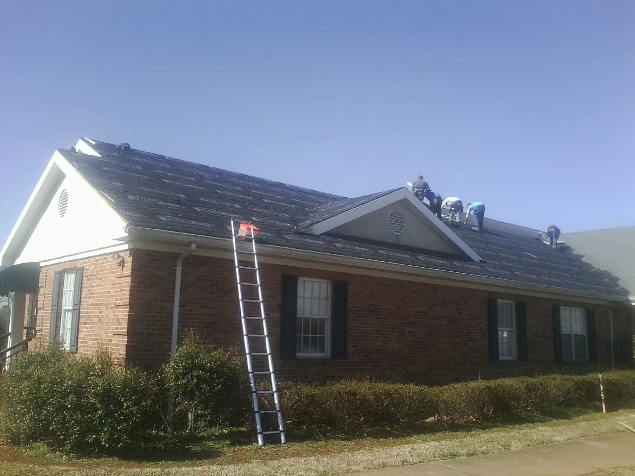 Crew tearing shingles off a brick ranch roof with a ladder set — Emergency Roof Repair work by our crew in Coon Rapids
