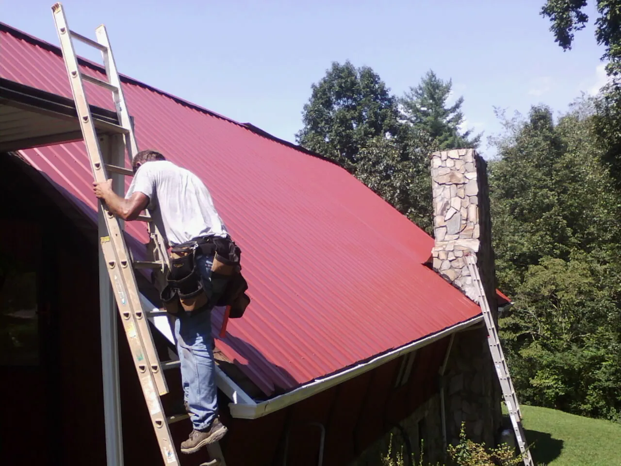 Roofer on a ladder beside a red metal roof and stone chimney — Soffit & Fascia Repair by Roofing Brothers in Coon Rapids
